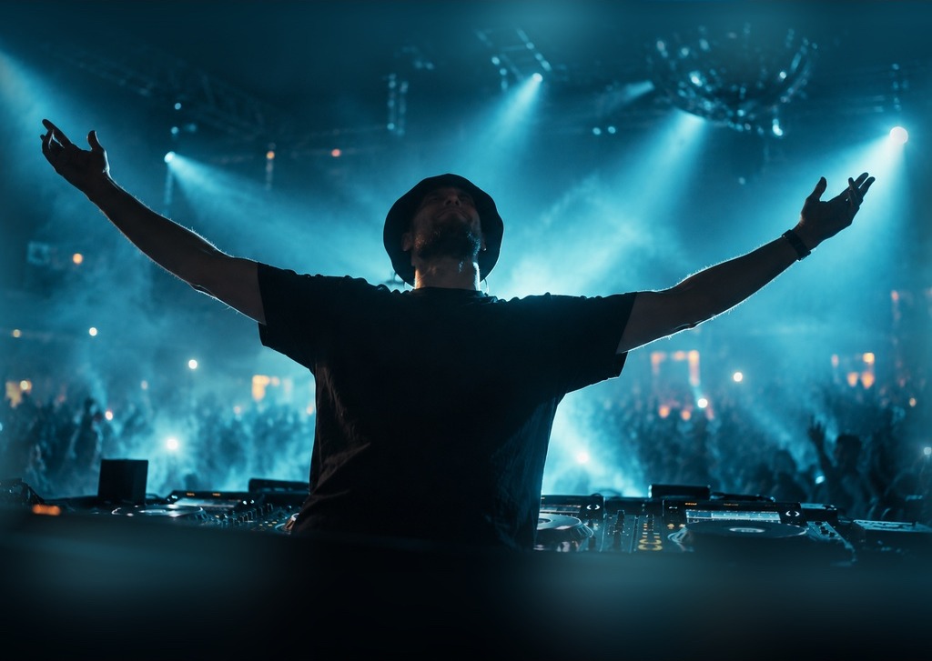 DJ in a black bucket hat with both hands raised amid teal-blue stage smoke